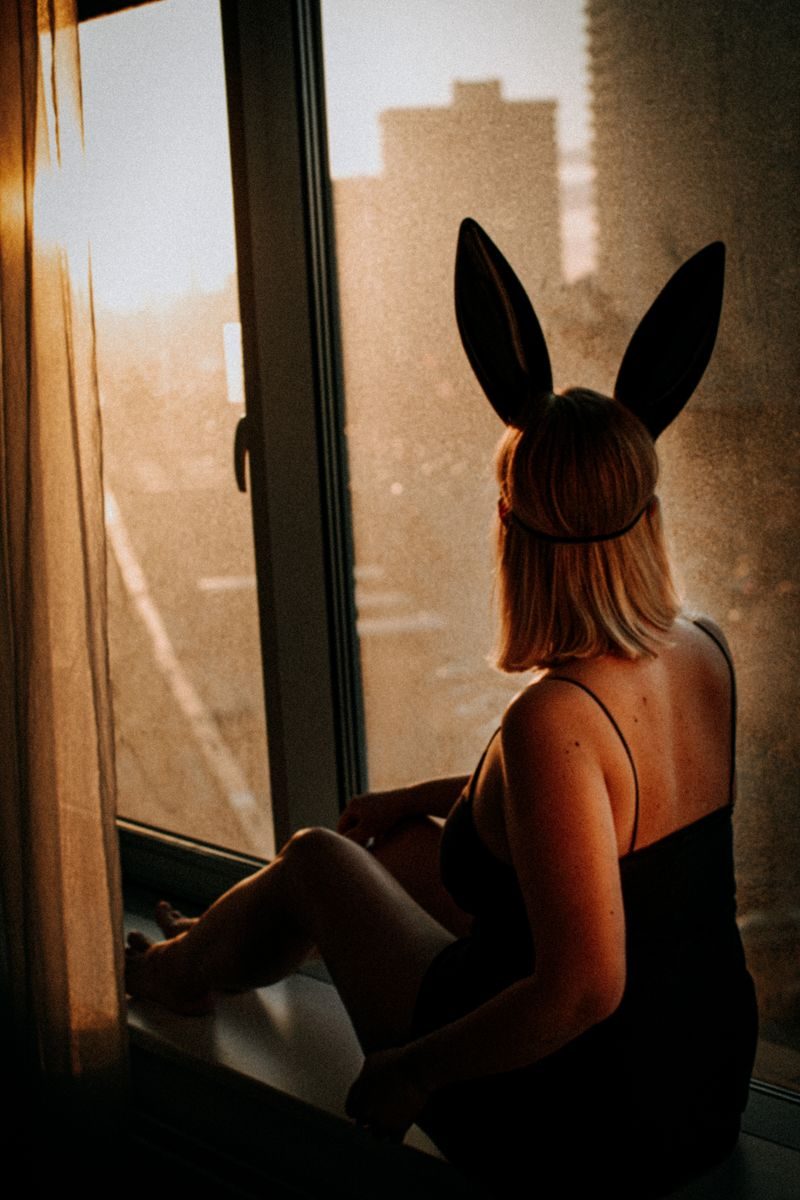 A woman with bunny ears sits by a window enjoying a sunrise view in the city.